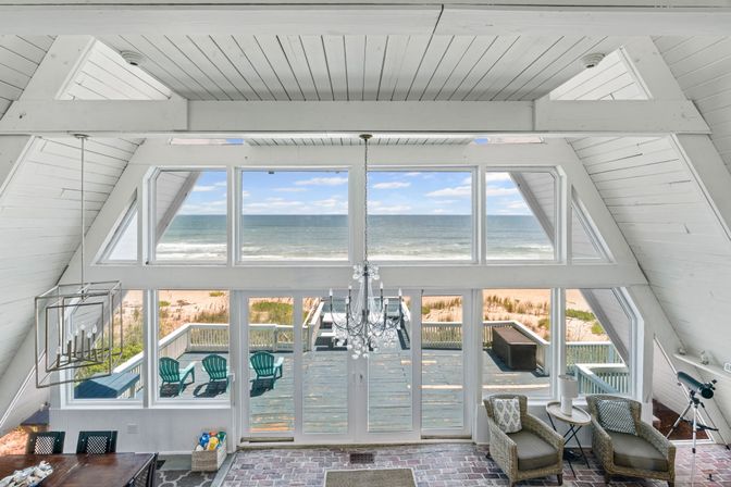 Bright A-frame beachfront living room with white shiplap ceiling, floor-to-ceiling windows and glass doors opening to a deck with teal Adirondack chairs, crystal chandelier, wicker armchairs and a telescope overlooking sandy beach and ocean.