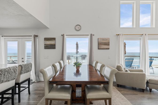 Bright beachfront dining room with long wooden table, eight upholstered chairs, small plant centerpiece and glass doors opening to oceanfront deck with lounge chairs and umbrella.