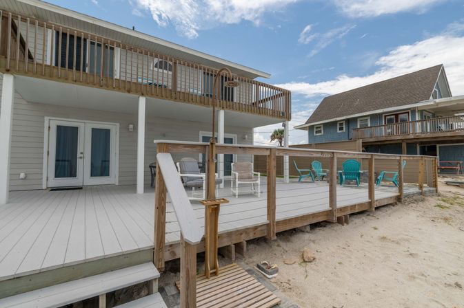 Oceanfront two-story beach house with wide wooden deck, cable-rail balustrade, turquoise Adirondack chairs, outdoor shower, sandy shore and upper balcony under a blue sky.