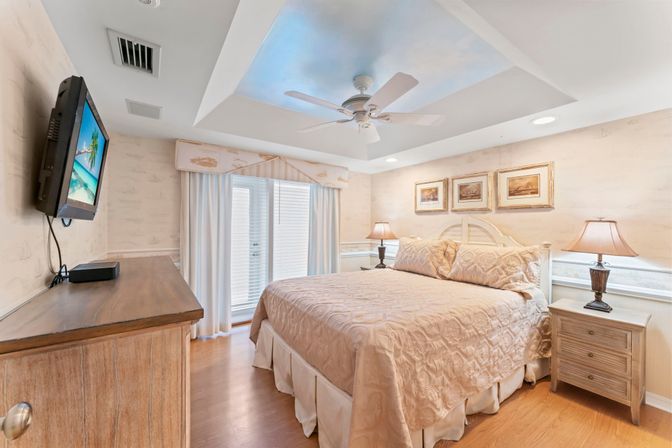 Coastal-inspired bedroom with a queen/king bed in beige quilt, ceiling fan, wall-mounted TV, hardwood floors, sliding glass doors with curtains, two nightstands with lamps and framed art