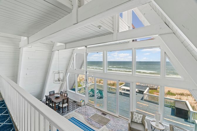 Sunlit white A-frame beach house interior with floor-to-ceiling windows framing ocean waves and sandy beach, open living and dining area with chandelier and seaside deck