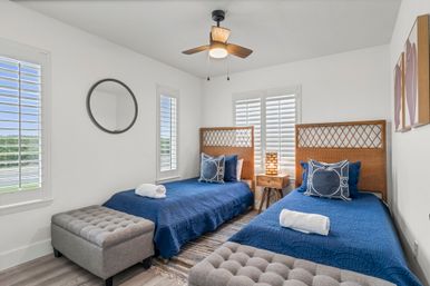 Bright coastal-style guest bedroom with two twin beds in navy quilts, woven rattan headboards, tufted gray ottomans, wooden bedside table with lamp, ceiling fan and white plantation shutters letting in natural light.