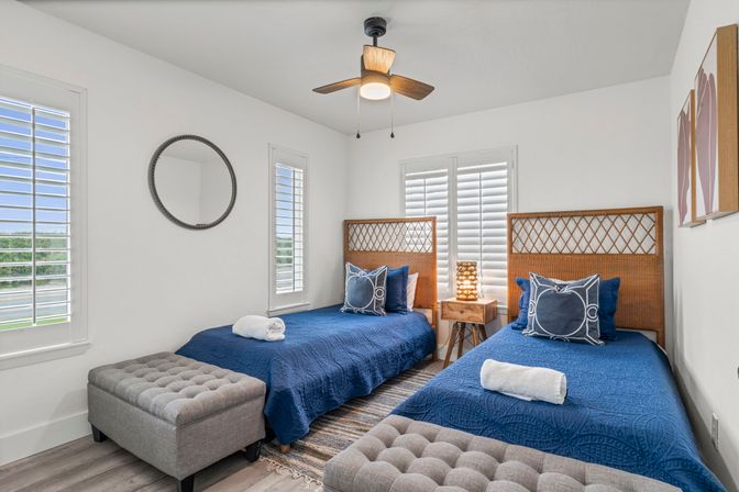 Bright coastal-style guest bedroom with two twin beds in navy quilts, woven rattan headboards, tufted gray ottomans, wooden bedside table with lamp, ceiling fan and white plantation shutters letting in natural light.