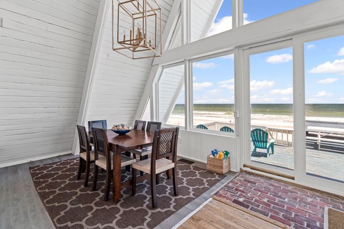 Sunlit beachfront A-frame dining room with a wooden six-seat table, patterned rug and geometric chandelier, large windows and sliding glass doors opening to a deck with teal Adirondack chairs and expansive ocean horizon.