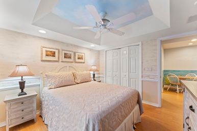 Coastal-style master bedroom with tray ceiling and ceiling fan, large bed with textured beige quilt, matching white nightstands and lamps, nautical prints above the headboard, hardwood floors and white closet doors.