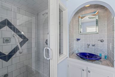 Bright bathroom with glass-enclosed tiled shower featuring a dark diamond accent and white grab bar, next to a recessed vanity with gray marble tiles, mirrored cabinet and a cobalt-blue glass vessel sink.