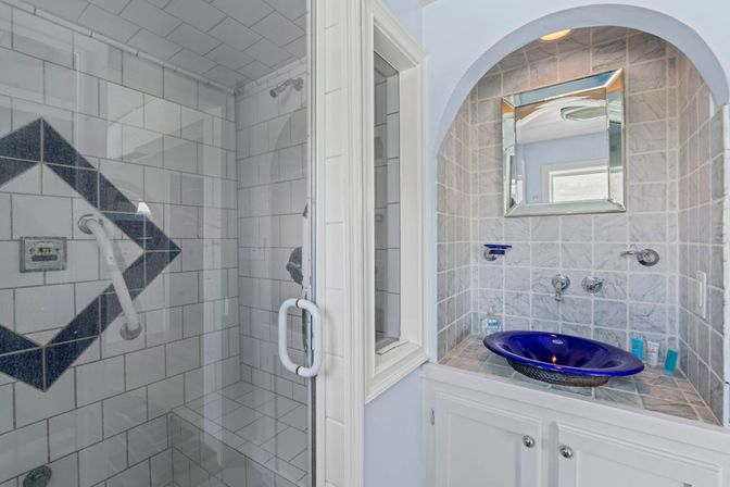 Bright bathroom with glass-enclosed tiled shower featuring a dark diamond accent and white grab bar, next to a recessed vanity with gray marble tiles, mirrored cabinet and a cobalt-blue glass vessel sink.