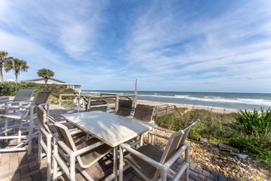 Oceanfront brick patio with outdoor dining table and lounge chairs, palm trees and a boardwalk leading to a sandy beach and rolling waves under a blue, wispy-cloud sky
