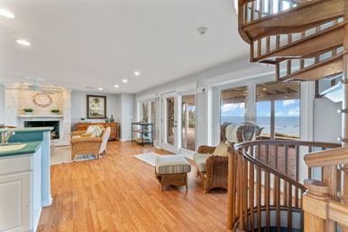 Bright coastal living room with hardwood floors, wicker armchairs and ottoman, stone fireplace, wooden spiral staircase, and ocean view through glass doors onto a beachfront patio.