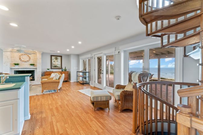 Bright coastal living room with hardwood floors, wicker armchairs and ottoman, stone fireplace, wooden spiral staircase, and ocean view through glass doors onto a beachfront patio.