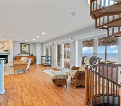 Bright coastal living room with hardwood floors, wicker armchairs and ottoman, stone fireplace, wooden spiral staircase, and ocean view through glass doors onto a beachfront patio.
