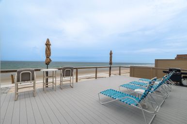 Oceanfront beachfront deck with blue woven lounge chairs, small table and chairs with closed umbrellas, gas grill and sandy beach meeting calm sea under a cloudy sky