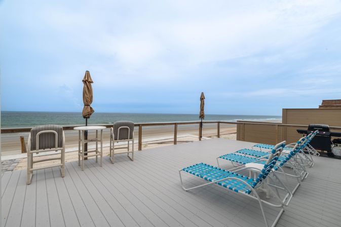 Oceanfront beachfront deck with blue woven lounge chairs, small table and chairs with closed umbrellas, gas grill and sandy beach meeting calm sea under a cloudy sky