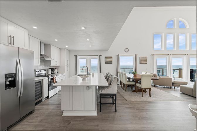 Sunlit open-concept coastal kitchen and living room with a white island, stainless-steel appliances, dining table, and large windows showcasing ocean and balcony views.