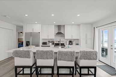 Sunlit modern white coastal kitchen with a large marble island, four cushioned barstools topped with gray faux-fur throws, stainless appliances and range hood, and French doors opening to an ocean view.