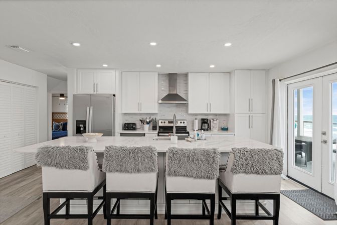 Sunlit modern white coastal kitchen with a large marble island, four cushioned barstools topped with gray faux-fur throws, stainless appliances and range hood, and French doors opening to an ocean view.