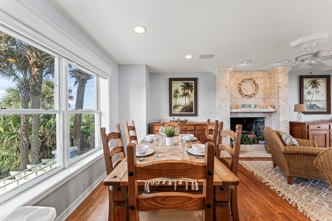 Bright coastal dining room in a beach house with a wooden farmhouse table set for a meal, large window framing palm trees, stone fireplace with wreath, wicker chairs and tropical artwork.