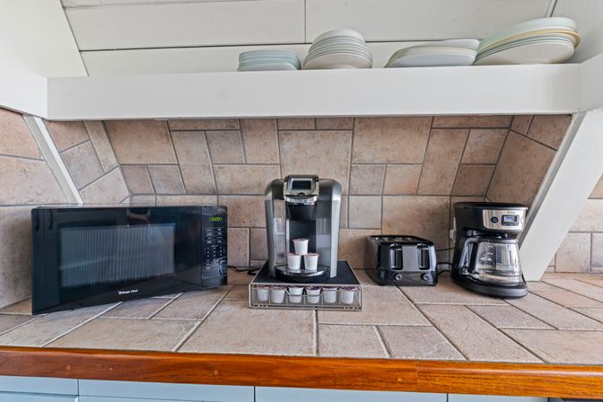 Tile-backed kitchen counter coffee station with a microwave, single-serve pod coffee maker and visible pods, two-slice toaster, and drip coffee pot under a shelf of stacked plates.
