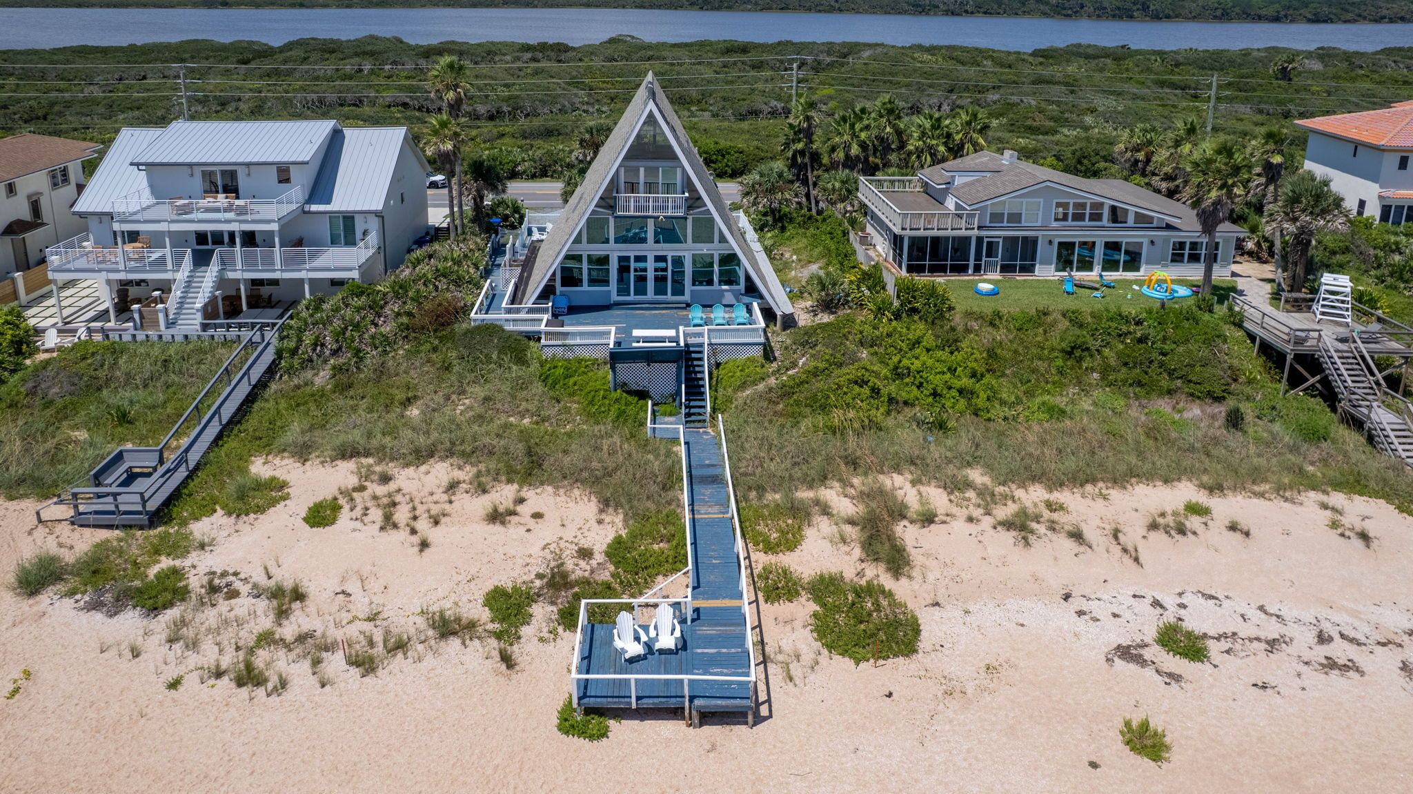 Oceanfront A-frame beach house with a long boardwalk to a sandy beachfront deck and Adirondack chairs, set among coastal dunes with neighboring beach homes and a lagoon in the background.