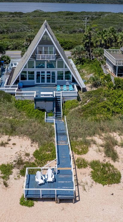 Oceanfront A-frame beach house with a long boardwalk to a sandy beachfront deck and Adirondack chairs, set among coastal dunes with neighboring beach homes and a lagoon in the background.
