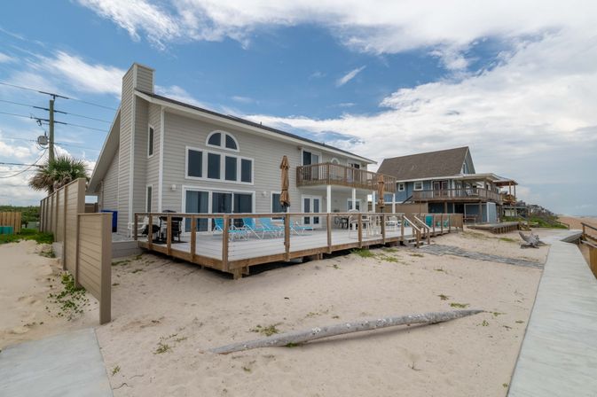 Oceanfront two-story beach house with large wooden deck, lounge chairs and umbrellas on a sandy shore beside a boardwalk and neighboring coastal homes under a partly cloudy sky.