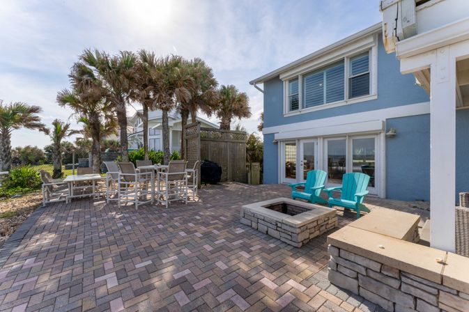 Coastal beachfront patio with brick pavers, palm trees, outdoor dining set, stone fire pit and turquoise Adirondack chairs beside a blue two-story beach house under a sunny sky