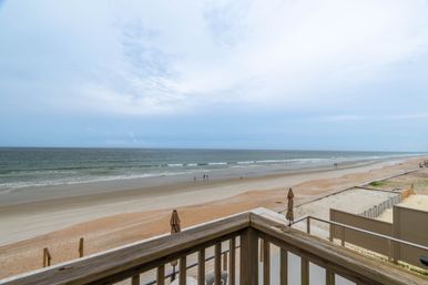 Oceanview from a wooden balcony overlooking a wide sandy beach and gentle waves under a pale cloudy sky, closed umbrellas and a few people strolling along the shoreline.