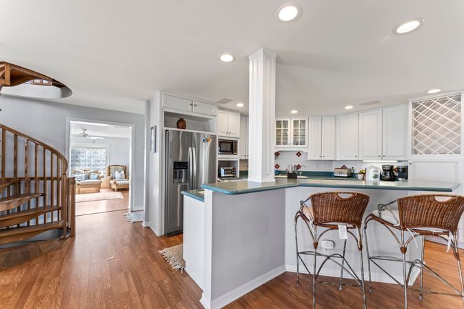 Bright open-concept kitchen with white cabinets, stainless steel refrigerator, teal countertop breakfast bar with wicker bar stools, hardwood floors and a wooden spiral staircase opening to a sunlit living room
