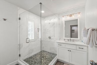 Bright modern bathroom with frameless glass walk-in shower, pebble-tile shower floor and marble-look walls, white vanity with lighted mirror and neatly folded towels