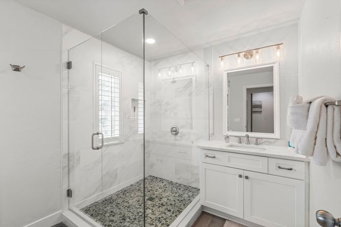 Bright modern bathroom with frameless glass walk-in shower, pebble-tile shower floor and marble-look walls, white vanity with lighted mirror and neatly folded towels