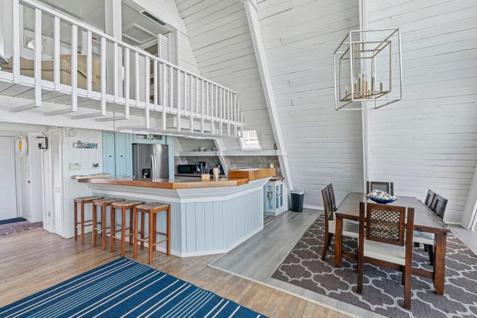Bright A-frame coastal cottage interior with open-plan kitchen and dining area, white shiplap walls and loft railing, light-blue breakfast bar with wooden stools and butcher-block countertop, stainless fridge, wooden dining table under a geometric pendant.