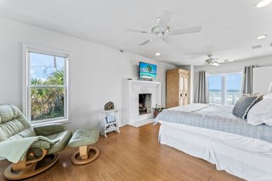 Sunny ocean-view coastal master bedroom with king bed, hardwood floors, white tiled fireplace and TV, green leather recliner with ottoman, and sliding glass doors to a balcony overlooking the sea.