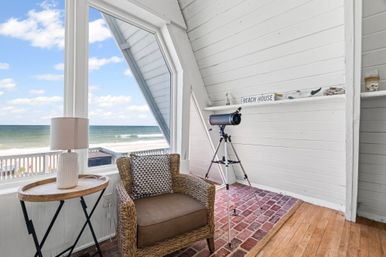 Cozy coastal reading nook in a beachfront A-frame cottage: wicker armchair with patterned pillow, round side table and lamp, telescope on a tripod by a large window with a sandy beach and ocean view under a blue sky.