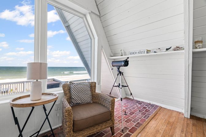 Cozy coastal reading nook in a beachfront A-frame cottage: wicker armchair with patterned pillow, round side table and lamp, telescope on a tripod by a large window with a sandy beach and ocean view under a blue sky.