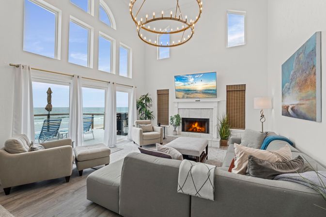 Bright beachfront coastal living room with two-story arched windows and sliding glass doors to an ocean-view deck with lounge chairs; neutral sectional and armchairs around a lit fireplace, wall-mounted TV, oversized circular chandelier, plants and beach-themed artwork.