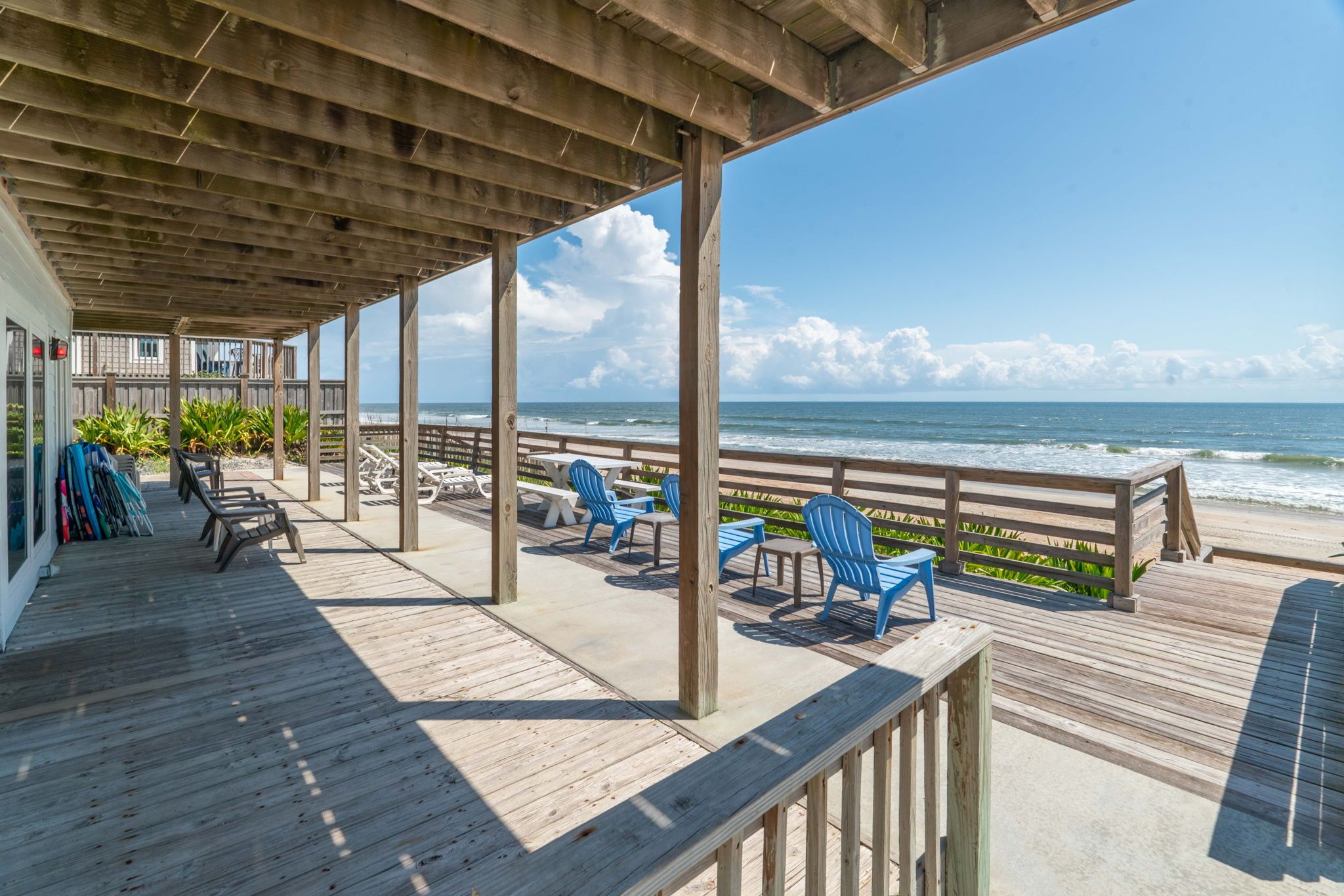 Sunny beachfront wooden deck and covered patio with blue Adirondack chairs and loungers facing the ocean, sandy beach and clear blue sky