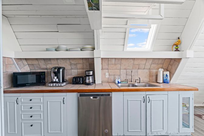 Sunlit coastal-style attic kitchenette with white shiplap ceiling and skylight, wood countertop, tile backsplash, double sink, stainless dishwasher and coffee station.