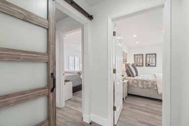 Bright modern hallway with a wood sliding barn door opening to two cozy bedrooms — one with plantation shutters and a twin bed, the other with patterned bedding, decorative mirrors and gray wood-look floors.