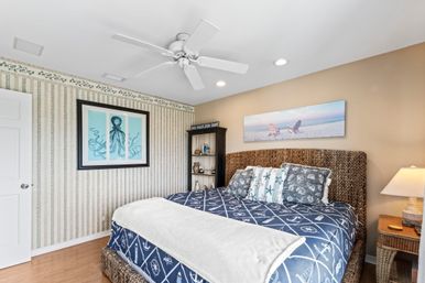 Coastal beach-house bedroom with king-size bed in navy nautical bedding, woven rattan headboard, starfish and seashell pillows, ocean and octopus wall art, ceiling fan and wicker nightstand with lamp
