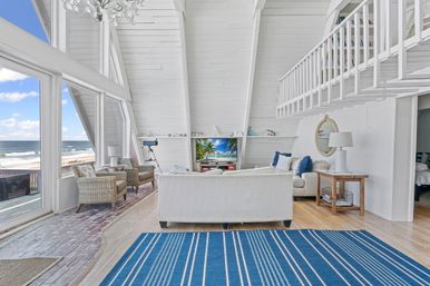Sunlit beachfront A-frame living room with vaulted white shiplap, floor-to-ceiling windows overlooking the ocean, wicker chairs and white sofas, blue striped rug, loft railing and telescope for coastal views