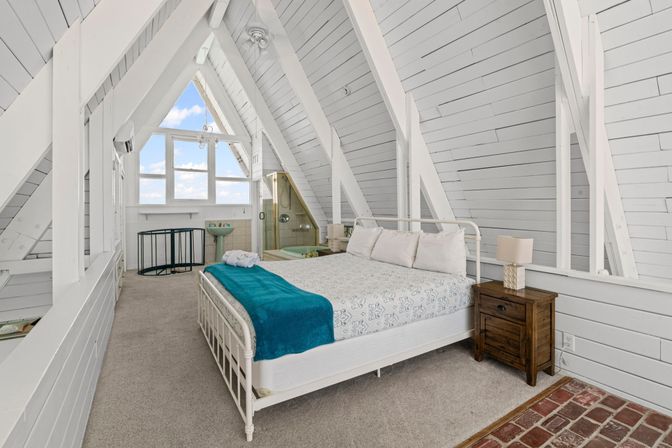 Cozy white A-frame loft bedroom with vaulted shiplap walls, queen bed topped with a teal throw, wooden nightstand, brick entry, and triangular window showing blue sky; small ensuite shower and pedestal sink.