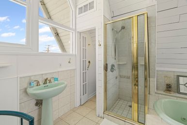 Bright beach-cottage bathroom with pastel green pedestal sink and tub, glass shower with brass trim, white shiplap walls, tile floor and large windows showing blue sky.