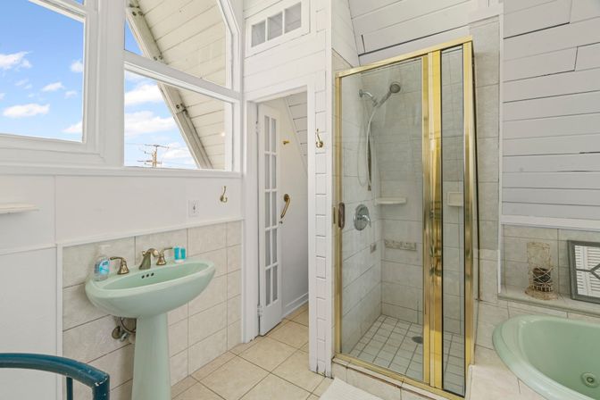 Bright beach-cottage bathroom with pastel green pedestal sink and tub, glass shower with brass trim, white shiplap walls, tile floor and large windows showing blue sky.