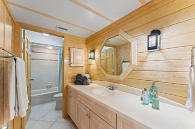 Wood-paneled bathroom with long white vanity, octagonal mirror, stacked gray towels, nautical glass bottles, lantern wall sconces, tile floor and bathtub/shower visible through an open door.