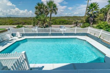 Sunlit turquoise swimming pool with white picket fence, Adirondack chairs and palm trees overlooking coastal dunes and a roadside