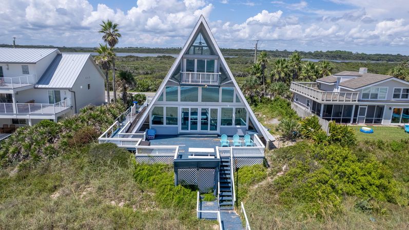 Beachfront A-frame house with large glass facade and wraparound deck, bright blue Adirondack chairs, wooden boardwalk over sandy dunes to the shore, palm trees and coastal marsh under a sunny sky.