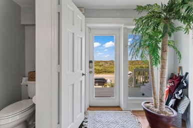 Bright beachside entryway with an open white door and glass door framing dunes and blue sky beyond; textured rug, large potted palm, umbrella stand, and a small half-bath visible to the left.