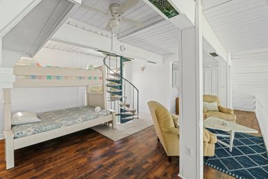 Bright coastal-style loft bedroom with white-painted beams and ceiling fan, metal spiral staircase, painted bunk bed, dark hardwood floors, and two tan armchairs with a white coffee table on a navy geometric rug.