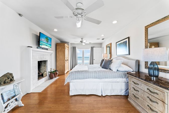 Bright beachfront master bedroom with coastal décor, king bed, ceiling fans, hardwood floors, fireplace, TV and balcony doors opening to ocean view