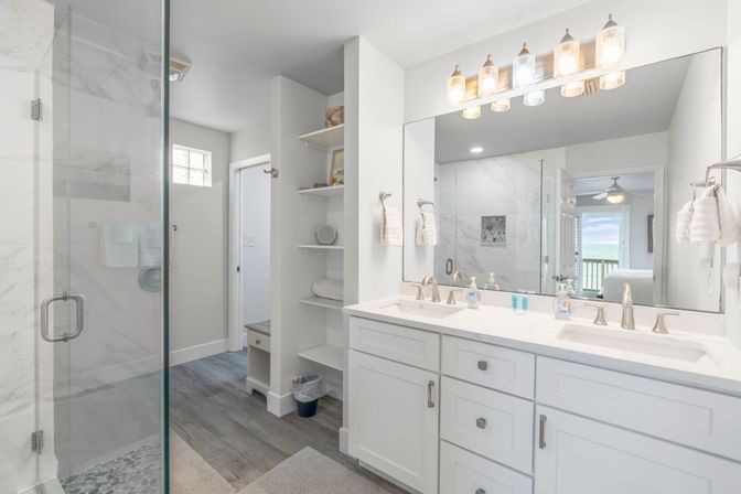 Bright coastal-style bathroom with double white vanity and brass faucets, large mirror reflecting a balcony ocean view, glass-enclosed marble shower, open shelving and wood-look floors.